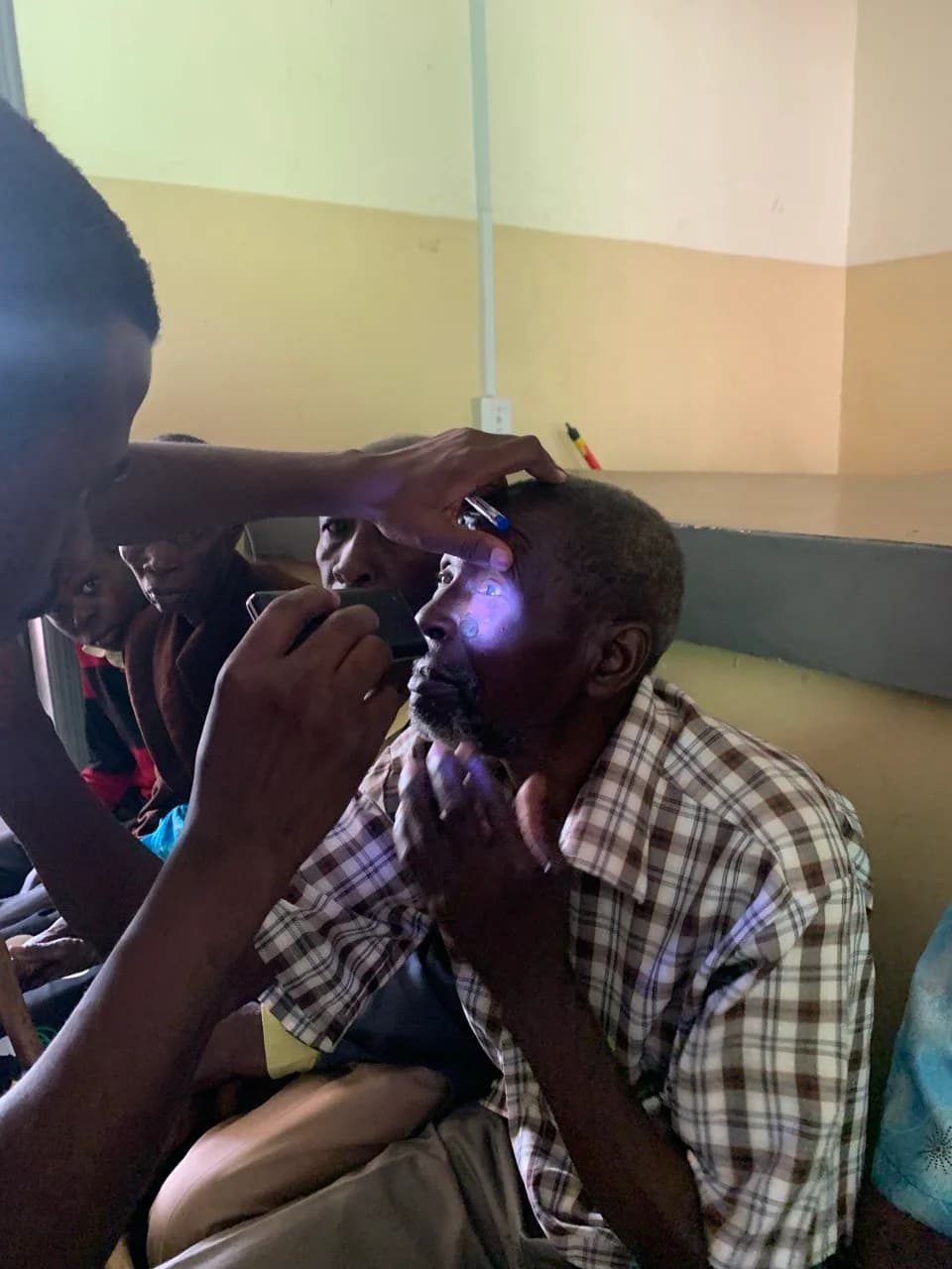 An older man receives an eye examination using a penlight, with other people in the room.